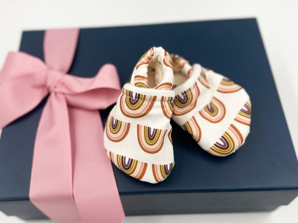 A pair of baby moccasins with a vibrant rainbow design, placed inside a box with a pink ribbon.